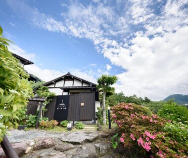 Stone path and flowering shrubs leading to traditional wooden gate of Yufuin Onsen Tsukanoma with distant mountain backdrop in Ōita