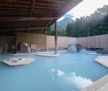 Outdoor milky-blue onsen pool with wooden roof, stone platforms and mountain backdrop