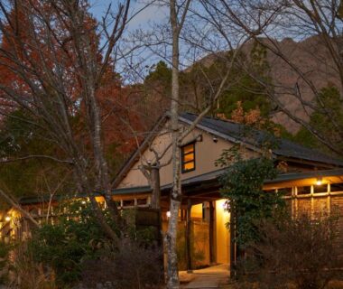 Twilight wooden ryokan entrance lit warmly, autumn trees and mountain backdrop at Yufuin Rakuyu, Ōita, Kyūshū