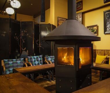 Lounge with central wood-burning stove, wooden benches and colorful cushions at Yufuin Tabinokura, Ōita