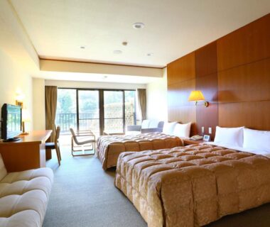 Bright Western-style twin room with two beds, sofa, balcony doors and wooden accent wall at Yugawara Chiyodasou, Kanagawa