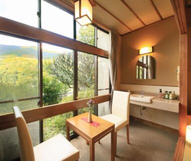 Seating area with two chairs, small table, vanity and large window framing green hills at Yugawara Ohnoya Ryokan Kanagawa
