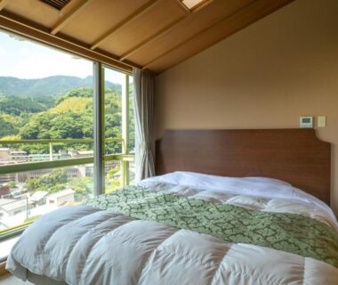 Bed beside large window with green mountain and town view, wood ceiling and tatami floor