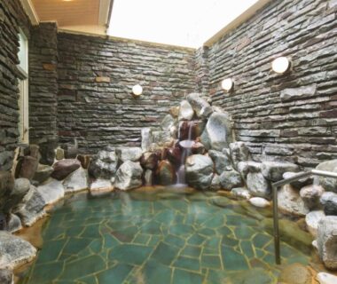Indoor stone-lined onsen bath with rock waterfall and skylight at Yugawara-Onsen Manyoso, Kanagawa