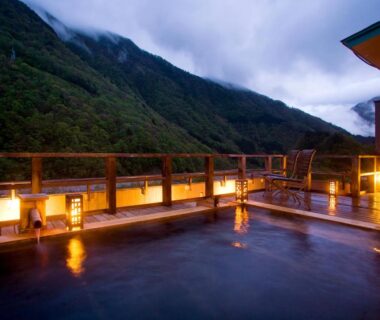 Outdoor onsen bath on wooden terrace with lantern lighting overlooking forested mountain valley