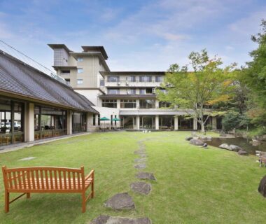Lawn courtyard with wooden bench, stepping stones and multi-story ryokan building surrounded by trees in Hyōgo