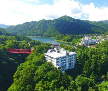 White ryokan building on forested hillside near red bridge, lake and green mountains in Gunma