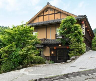Exterior of a traditional wooden ryokan with lantern-lit entrance and lush greenery on a sloped street in Oita