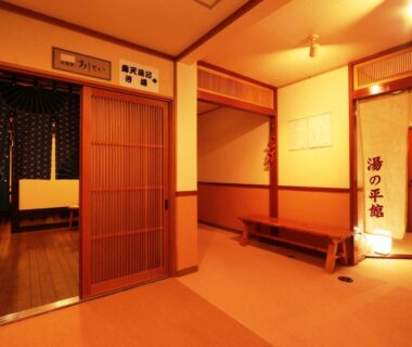 Entrance corridor with wooden sliding doors, bench, noren curtain and warm lantern lighting at Yunohirakan in Gifu