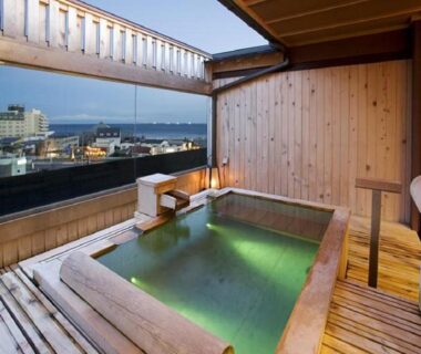 Outdoor wooden onsen bath on balcony overlooking waterfront and urban skyline at Yunokawa Kanko Hotel Shoen