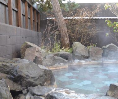 Steaming rock-lined outdoor onsen with wooden fence, stone edging, and a tree at Yunokawa Onsen Emi Hakodateya, Hokkaidō