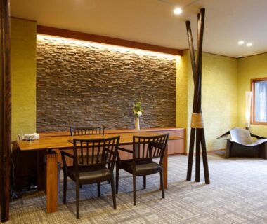 Interior lounge with wooden desk, chairs, textured stone wall and window at Yunokawa Onsen Emi Hakodateya Hokkaidō