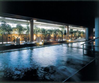Spacious indoor onsen bath with wooden trim, lanterns and potted plants lining a glass window at dusk