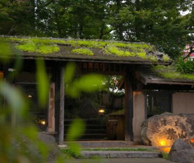 Traditional wooden gate with moss-covered roof and lit stone lantern at Yunosato Hayama in Oita