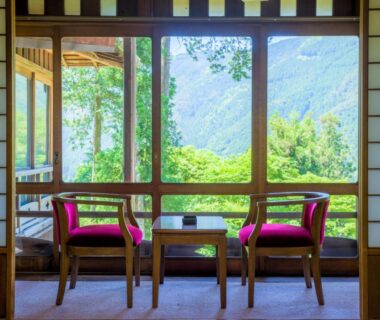 Sitting area with wooden chairs, low table and shoji screens overlooking forested mountains at Yunoshimakan in Gifu