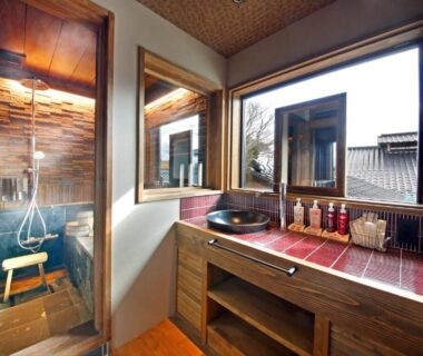 Compact wooden bathing area with tiled tub, handheld shower, red-tiled vanity, vessel sink and window overlooking rooftops