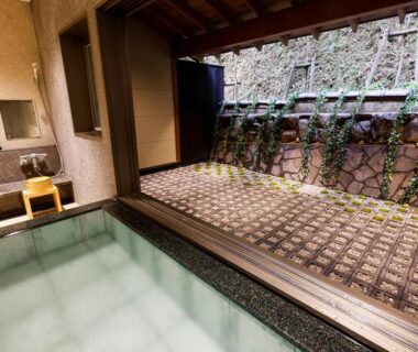 Indoor onsen bath with wooden deck, tiled tub and stone courtyard wall with climbing plants at Yuraku Kinosaki, Hyōgo