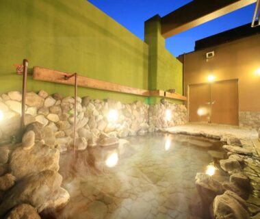 Stone-lined outdoor onsen with warm lantern lighting, stone wall and wooden rail at Yutorelo Beppu, Ōita