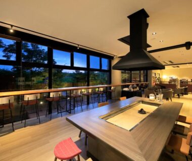 Spacious wooden dining area with central grill, bar seating and large windows overlooking dusk landscape in Kumamoto