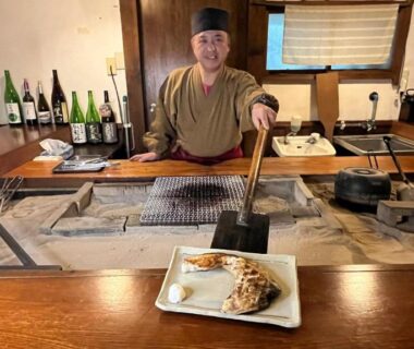 Staff cooking at irori hearth behind wooden counter with sake bottles, Yuuzuru ryokan in Shizuoka, Chūbu
