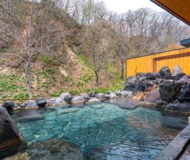 Rock-lined outdoor onsen bath at Yuyado DAIICHI beside wooden ryokan building with mountain backdrop in Hokkaidō