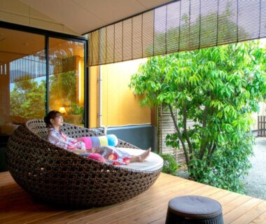 Guest reclining on wicker daybed on wooden terrace overlooking a small Japanese garden at Yuyado Ichibanchi, Shizuoka