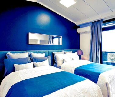 Bright twin bedroom with blue accent wall, two bedside lamps, balcony sliding door and white bedding with blue runners