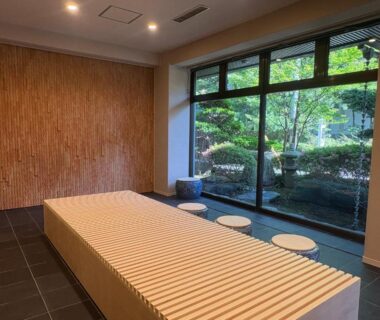 Minimal wooden bench and stools by large window overlooking lush courtyard garden at soraichi ryokan in Miyagi