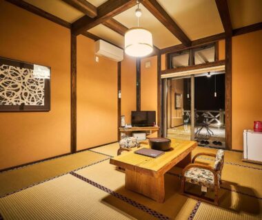 Tatami room with low wooden table, two cushioned floor chairs, TV, small fridge and sliding glass doors to balcony