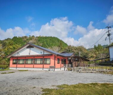 Single-story ryokan with orange facade, gravel courtyard and hillside backdrop at Chalet de montagne Hibari d’Asagiri, Kumamoto