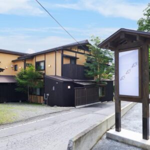 Wooden sign and two-story ryokan beside a gravel driveway and trees in Gunma, Kantō
