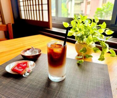 Glass of iced coffee with snacks on wooden table beside shoji screens and potted plant in Ryokan Uribō, Gunma