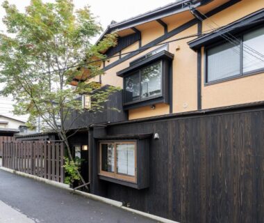 Exterior of Ryokan Uribō with dark wooden fence, beige walls and entrance along narrow street in Gunma