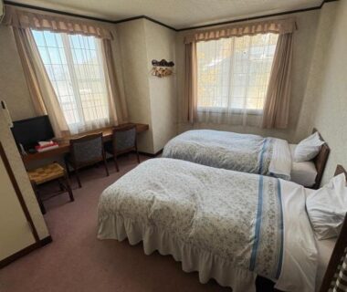 Twin beds with decorative bedcovers, small desk, TV and two curtained windows in Oyado Marutoya room Nagano