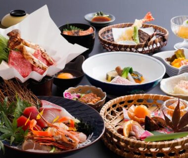 Assorted small Japanese dishes in bowls and woven baskets, featuring prawns and grilled items on a black tray