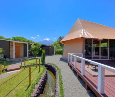 Tent-style lodge with wooden deck, gravel path and small cabins in grassy courtyard with mountain backdrop, Yamanashi