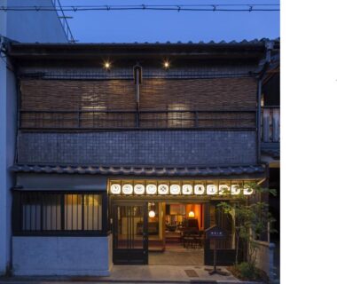 Two-story ryokan facade with lit lanterns, open wooden entrance and warm interior light at Dozen Ryokan & Cafe, Kyoto