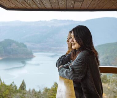 Two guests on a wooden balcony at Magohachi overlooking a lake and forested hills in Gifu