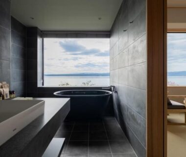 Dark tiled bathroom with freestanding stone tub and large window overlooking sea, adjacent tatami seating area visible