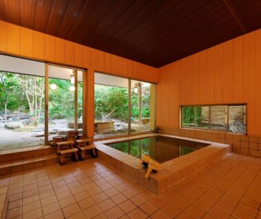 Indoor wooden onsen bath with tiled floor and garden views through large windows at Sanpei-sō, Shizuoka