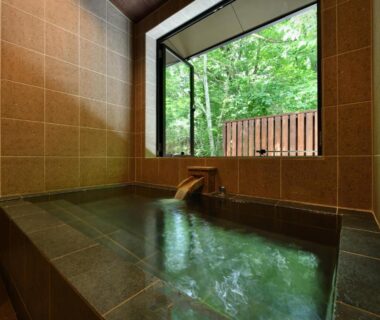 Tiled indoor onsen bath with wooden spout and large window framing green forest at Hakkaisan Konohanakan Yamanashi