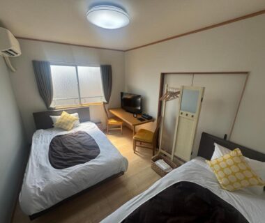 Compact twin room with two beds, TV, wooden floor, air conditioner and window at Manrikiya Bekkan Beppu