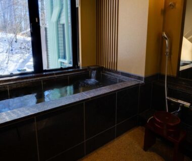 Indoor stone onsen bath with shower and wooden stool, window overlooking snowy trees at Bettei Tateshina Kunpū