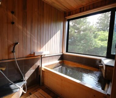 Wooden soaking tub and shower area with window overlooking greenery at Jinpyōkaku Honten, Nagano