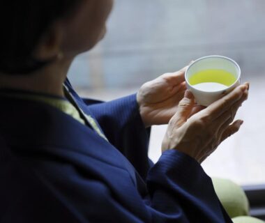 Guest wearing a dark robe holding a cup of green tea by a window at Hôtel touristique Tōnomine in Nara