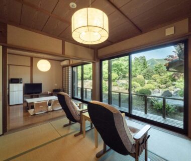 Traditional tatami room with low table, lounge chairs and sliding glass doors overlooking a manicured garden at Sansō Hanayu, Kumamoto