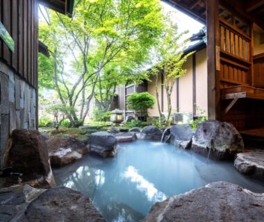 Rock-lined outdoor onsen bath surrounded by garden and wooden architecture at Sansō Hanayu Kumamoto