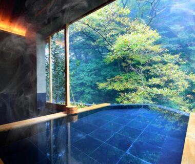 Steaming indoor onsen with stone bath and wooden frame facing lush green forest at Okuyugawara YUI Kanagawa