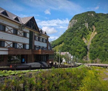 Multi-story ryokan building by a river with mountain backdrop, green foliage and small bridge in Sōunkyō Onsen Hokkaidō