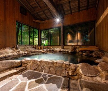 Wooden interior with rock-lined onsen bath and greenery visible through windows at Kosugian, Yamakawa Onsen, Kumamoto